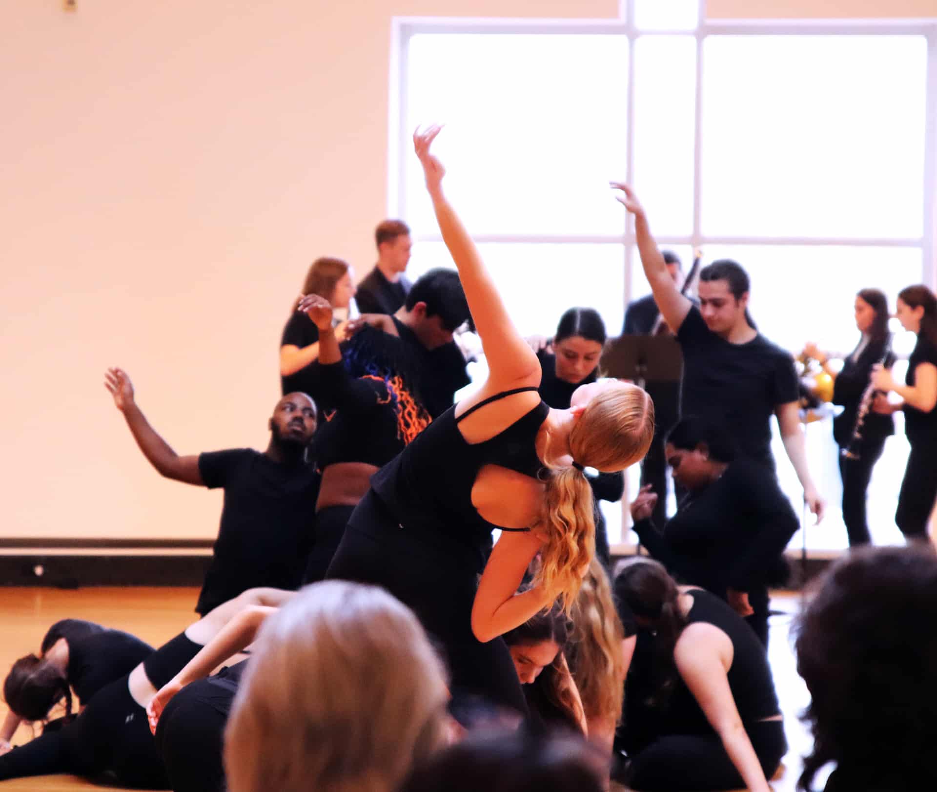 A group of dancers in black attire perform at the Harn Museum. In the center, a woman with long blonde hair arches her back, one arm raised, over a cluster of other dancers on the floor. In the background, other performers and musicians are visible in front of a large, bright window.